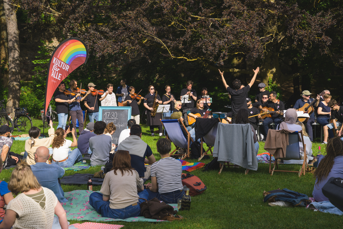 Ein Orchester auf spielt einer grünen Wiese, davor sitzen Zuhörer im Gras