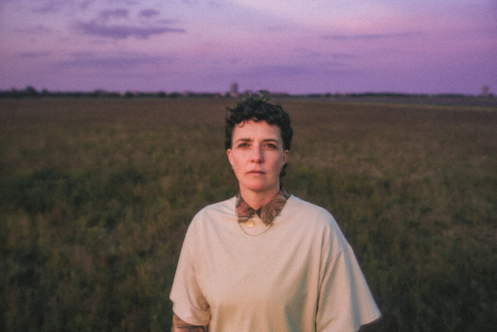 Junge Frau mit kurzen Haaren in einem hellen Pullover vor einem Feld. Violetter Himmel.
